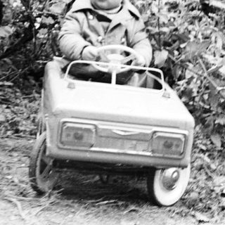 Here's a brief summary of the image: Child sits in a toy car outdoors, near foliage.