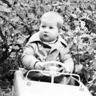 Here's a brief summary of the image: Child sits in a toy car outdoors, near foliage.