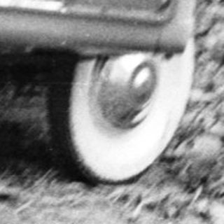 Here's a brief summary of the image: Child sits in a toy car outdoors, near foliage.