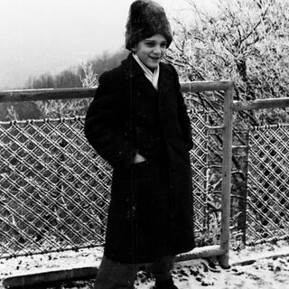 Young person in winter coat and hat, standing near a fence.