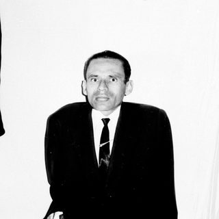 Man in suit stands against a wall with bookshelves.