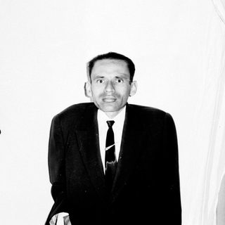 Man in suit stands against a wall with bookshelves.