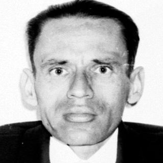 Man in suit stands near a bookshelf, black and white photo.