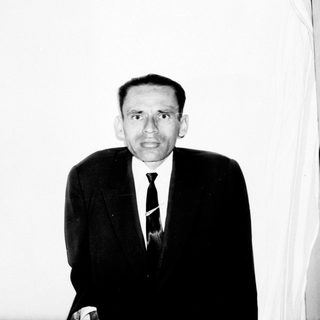 Man in suit stands near a bookshelf, black and white photo.