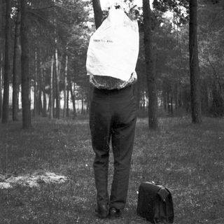 Man with bag over head stands in a wooded area.