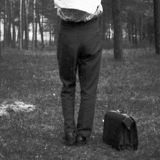 Man with bag over head stands in a wooded area.