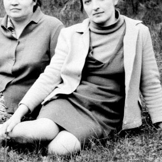 Two women sit in grass, surrounded by trees.