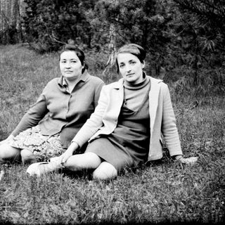 Two women sit in grass, surrounded by trees.
