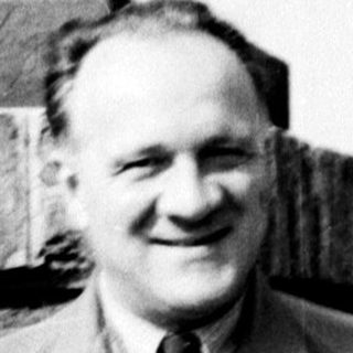 Man in coat stands near brick wall, smiling.