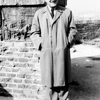 Man in coat stands near brick wall, smiling.