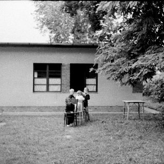 People seated outdoors near a building, shaded by a tree.