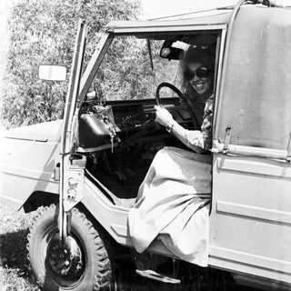 Woman in vintage vehicle, driving outdoors in a grassy area.