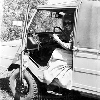 Woman in vintage vehicle, driving outdoors in a grassy area.