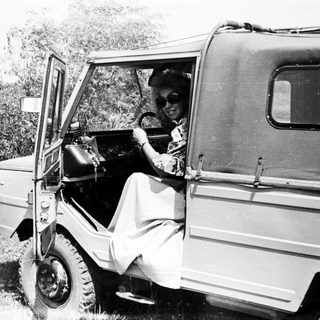 Woman driving a vintage vehicle in a grassy landscape.
