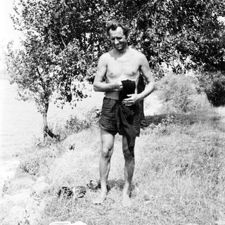 Man stands near water, partially clothed in a grassy area.