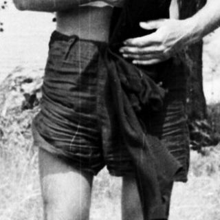 Man stands near water, partially clothed in a grassy area.