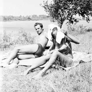 Two nude figures relax on a blanket in grassy field.