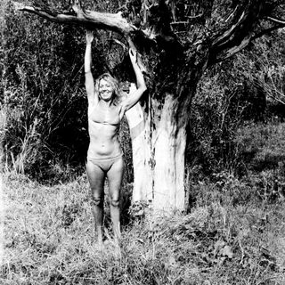 Nude person hangs from a tree branch in a field.