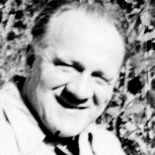 Man sitting outdoors, smiling, surrounded by vegetation.