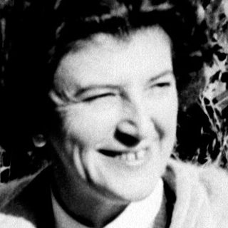 A woman with a camera smiles amidst tall grass.
