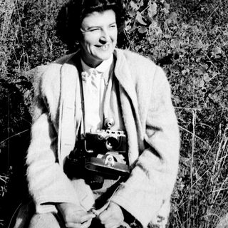 A woman with a camera smiles amidst tall grass.