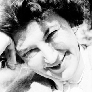Here's a brief summary of the image: Woman with curly hair smiles, posing outdoors with a hand on head.