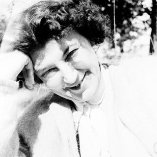 Here's a brief summary of the image: Woman with curly hair smiles, posing outdoors with a hand on head.