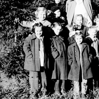 Group of children and an adult stand in a wooded area.