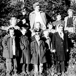 Group of children and an adult stand in a wooded area.