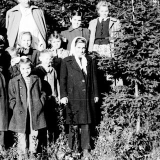 Group of children and an adult stand in a wooded area.