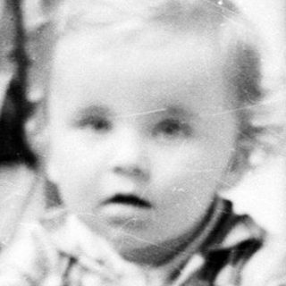 Here's a brief summary of the image: Baby stands near a railing, looking toward the camera.