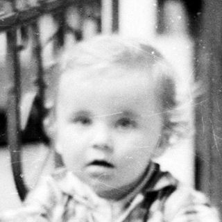 Here's a brief summary of the image: Baby stands near a railing, looking toward the camera.