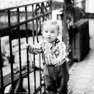 Here's a brief summary of the image: Baby stands near a railing, looking toward the camera.