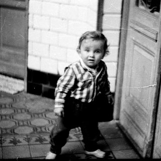 Baby stands near a door, looking directly at the camera.