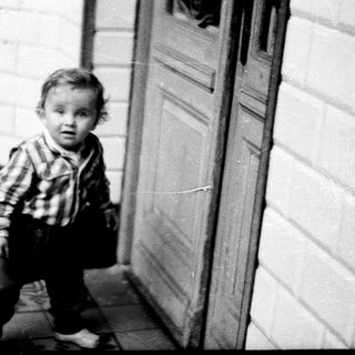 Baby stands near a door, looking directly at the camera.