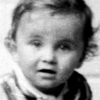 Baby stands near a door, looking directly at the camera.