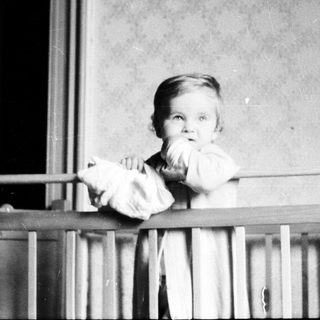 Baby peering over a crib railing in a vintage photograph.