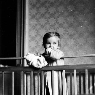 Here's a brief summary of the image: Baby standing in a crib, holding a doll.