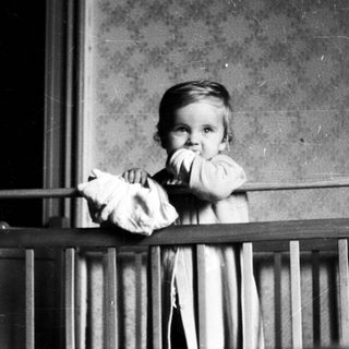Here's a brief summary of the image: Baby standing in a crib, holding a doll.