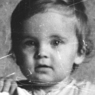 Here's a brief summary of the image: Baby stands behind a railing, looking directly at the camera.