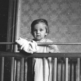 Here's a brief summary of the image: Baby stands behind a railing, looking directly at the camera.