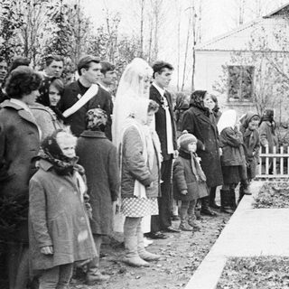 Group gathers as a man speaks outdoors, likely at a memorial.