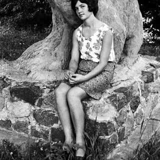 Here's a brief summary of the image: Woman seated near a stone wall and tree roots.