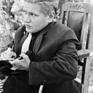 Man in suit sits outdoors, holding a cigarette.