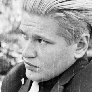 Man in suit sits outdoors, holding a cigarette.