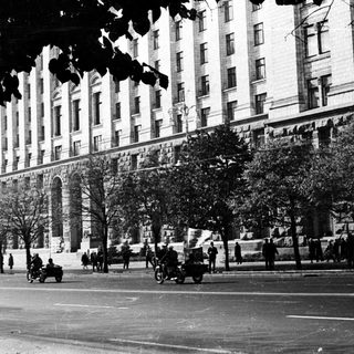 Here's a brief summary of the image: Black and white street scene with a large building and pedestrians.