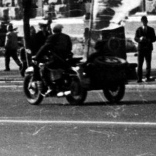Here's a brief summary of the image: Black and white street scene with a large building and pedestrians.