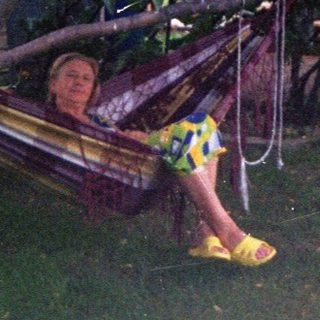 Person relaxes in a hammock under a tree, near a house.