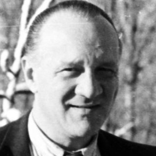 Man in suit stands outdoors with snow-covered trees.