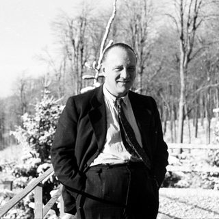 Man in suit stands outdoors with snow-covered trees.